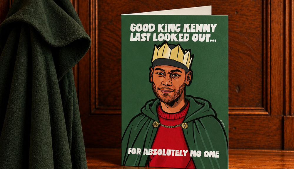 King Kenny Christmas card standing slightly open on a warm wooden mantelpiece beside a hanging green Traitor's cloak.