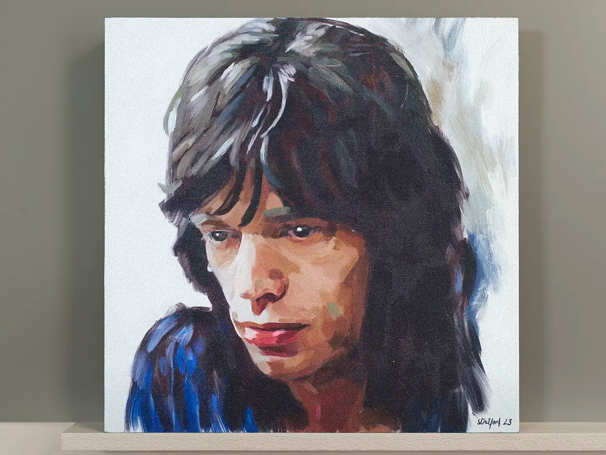 A square canvas painting of Mick Jagger with dark hair, shown leaning against a neutral wall on a light wooden surface.