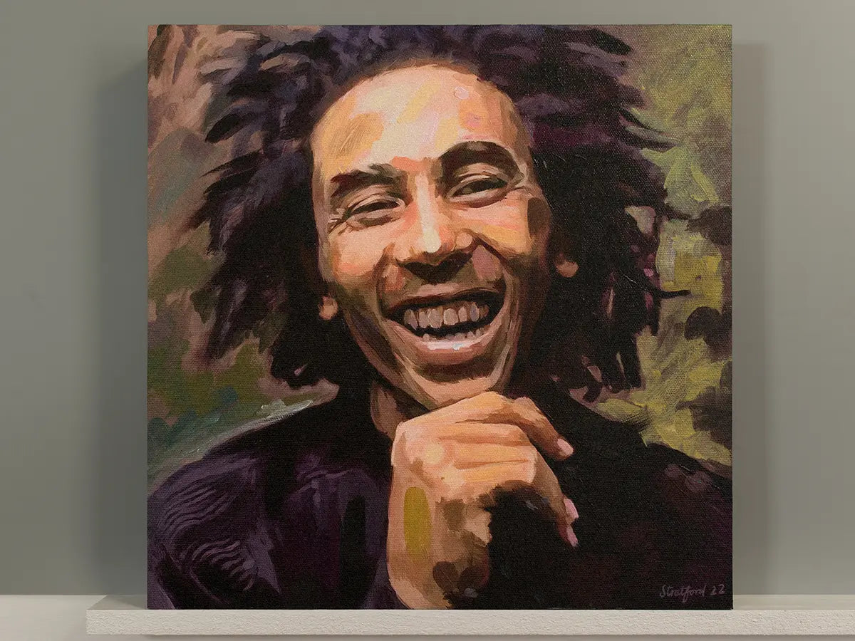 A square canvas painting of a smiling Bob Marley portrait, displayed against a neutral gray wall on a white shelf. The painting features expressive brushstrokes in dark purple, black, olive green, and warm skin tones.