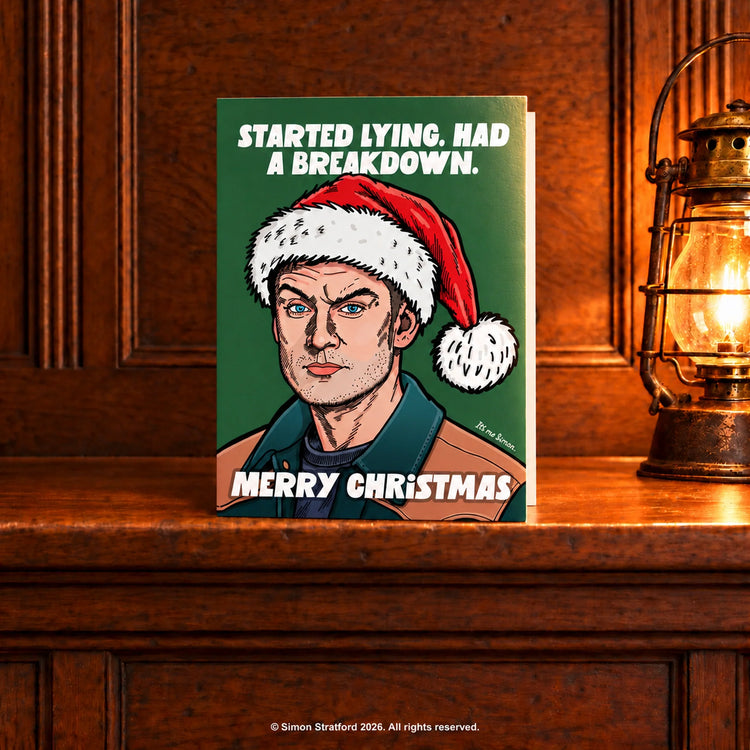 James Acaster Christmas card in a Santa hat: Started lying. Had a breakdown. Merry Christmas. Standing slightly open on a wooden mantelpiece beside a glowing antique lamp.
