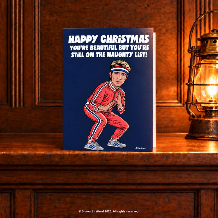 James Blunt Christmas card showing him in a red tracksuit: You’re beautiful but you’re still on the naughty list. Standing slightly open on a wooden mantelpiece beside a glowing antique lamp.