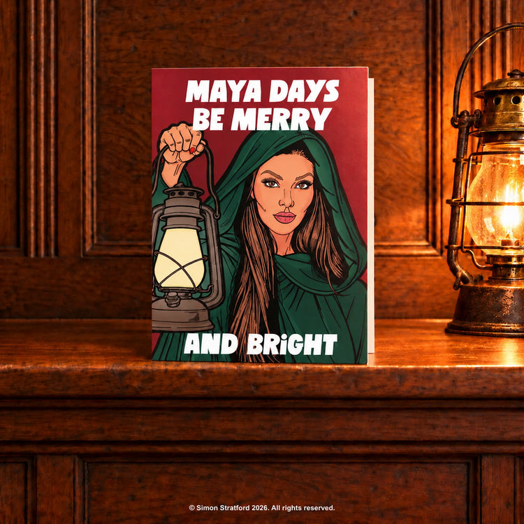 Maya Jama Christmas card showing her in a green cloak holding a lantern: Maya days be merry and bright. Standing slightly open on a wooden mantelpiece beside a glowing antique lamp.