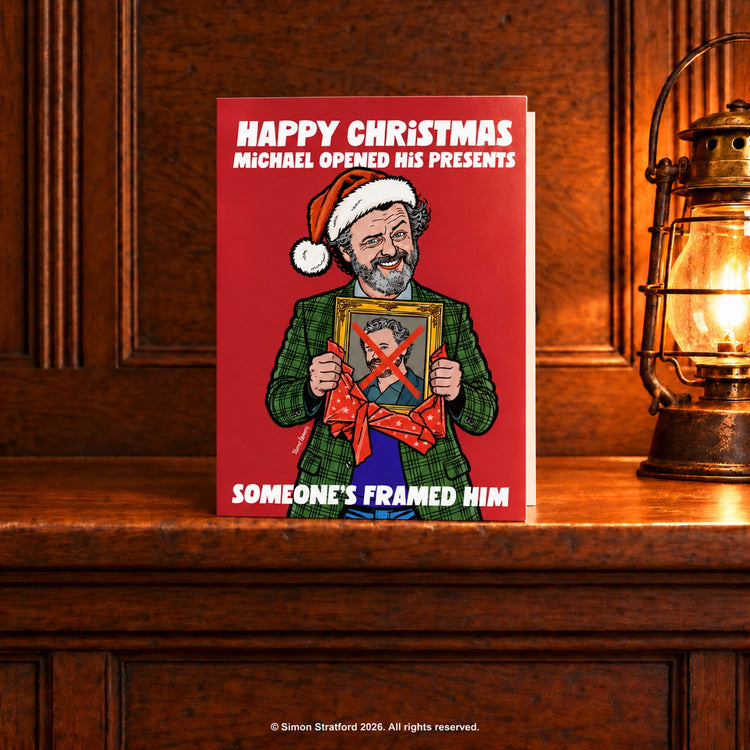 Michael Sheen Christmas card showing him in a Santa hat holding a framed portrait with a red X: Someone’s framed him. Standing slightly open on a wooden mantelpiece beside a glowing antique lamp.