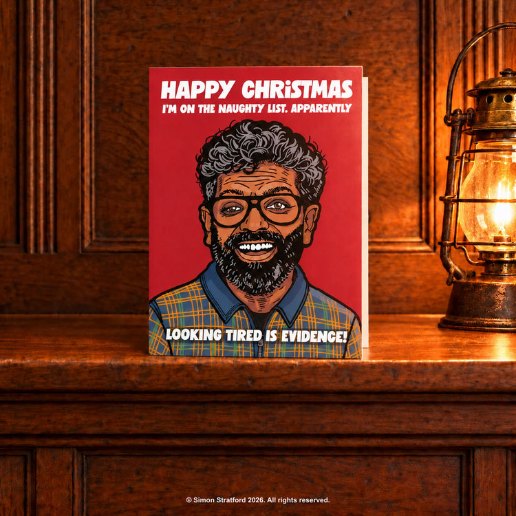 Romesh Ranganathan Christmas card with a red background: I’m on the naughty list. Apparently, looking tired is evidence. Standing slightly open on a wooden mantelpiece beside a glowing antique lamp.