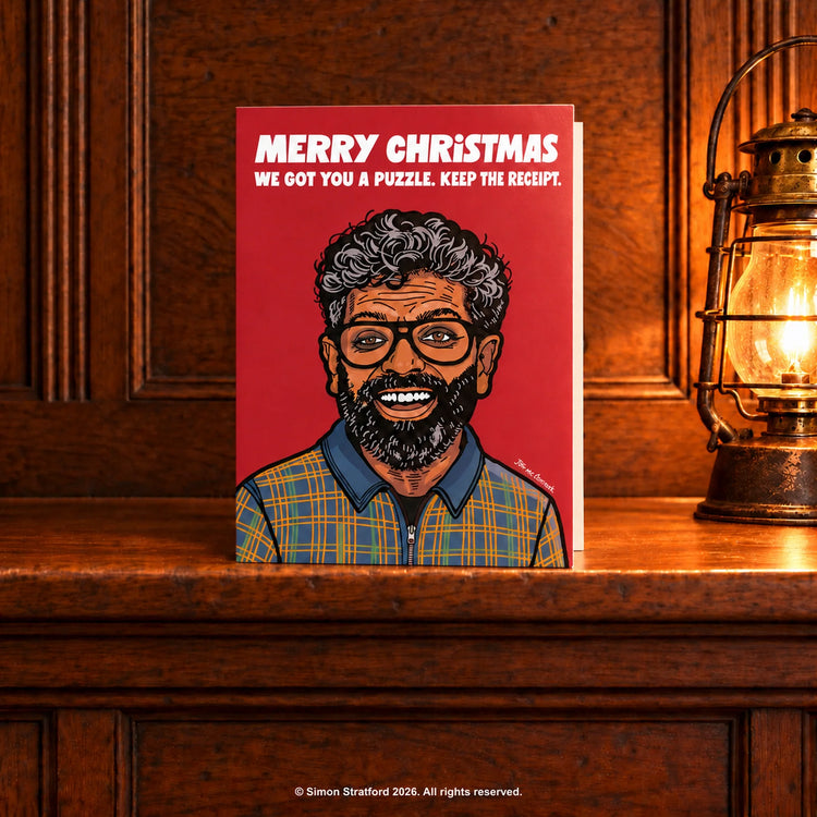 Romesh Ranganathan Christmas card: Merry Christmas. We got you a puzzle. Keep the receipt. Standing slightly open on a wooden mantelpiece beside a glowing antique lamp.